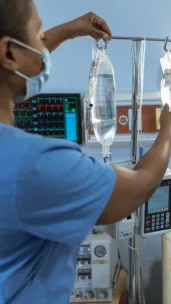 A healthcare worker wearing blue scrubs and a medical mask adjusts intravenous (IV) medicine hanging on a metal pole in a hospital room, with a patient monitor and hospital bed visible in the background. This image represents clinical research.